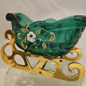 May include: A decorative glass sleigh in emerald green with gold accents. The sleigh features a floral design with white and red flowers. The base is gold-colored metal. A festive holiday decoration.