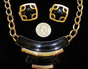 May include: A gold chain necklace with a black and gold geometric pendant. The necklace is paired with matching black and gold square earrings.