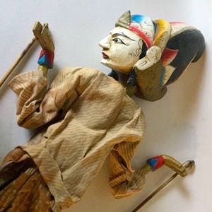 May include: A wooden puppet with a painted face and elaborate headdress. The puppet is wearing a brown and white striped shirt and brown pants. The puppet is holding two wooden sticks.