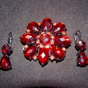 May include: A jewelry set consisting of a circular brooch and two matching drop earrings. The pieces feature faceted, deep red, pear-shaped stones arranged in a floral pattern, accented with smaller orange crystals set in dark metal frames.