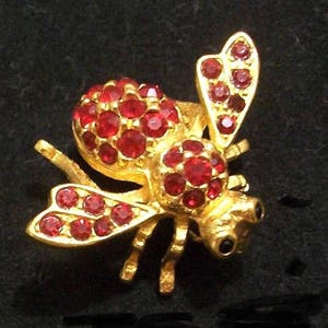 May include: A gold-toned bee brooch featuring a textured metal body adorned with numerous small, round, vibrant red crystals embedded across the wings, thorax, and abdomen, with two small black eyes visible on the head of the insect figure.
