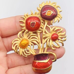 May include: Gold-tone brooch featuring a bouquet of three enamel sunflowers in a red and purple enamel vase.