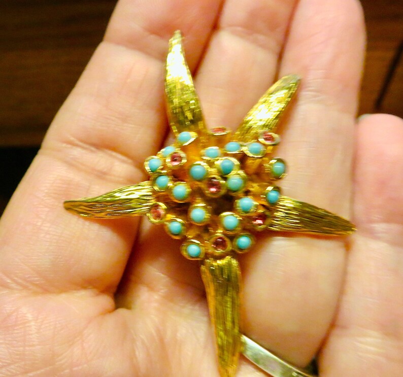 May include: A gold-toned starfish-shaped brooch with textured details. The center features clusters of turquoise and pink gemstones. The brooch is held in a hand, showcasing its size and design.