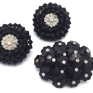 Peut inclure: Deux broches circulaires en perles noires avec un motif floral central argenté orné de strass transparents, et une broche ovale noire plus grande composée de multiples petites formes de fleurs, chacune accentuée par un strass central.