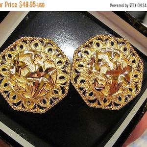 May include: A pair of gold-toned earrings with a filigree design. Each earring features a central starburst design surrounded by an ornate, openwork border. The earrings are presented in a black jewelry box.