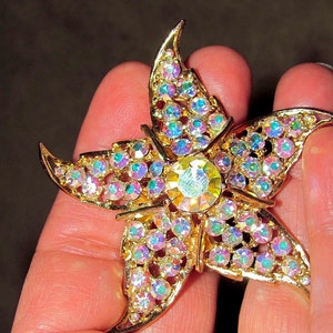 May include: A gold-tone starfish brooch featuring a large central faceted crystal surrounded by numerous smaller iridescent rhinestones. The metal frame has a textured finish, and the stones exhibit a multi-colored shimmer across the five arms.