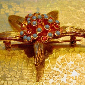 May include: A gold-toned starfish brooch with textured details. The center features a cluster of light blue and red-orange beads. The brooch has a pin closure on the back.