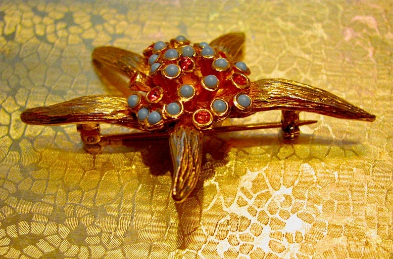 May include: A gold-toned starfish brooch with textured details. The center features a cluster of light blue and red-orange beads. The brooch has a pin closure on the back.