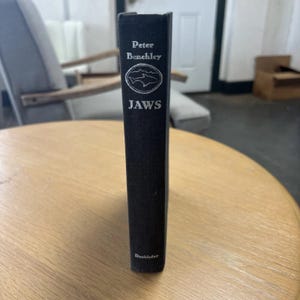 May include: A hardcover book titled "JAWS" by Peter Benchley. The book has a black cover with silver lettering and a circular graphic of a shark. The publisher's name, Doubleday, is at the bottom.