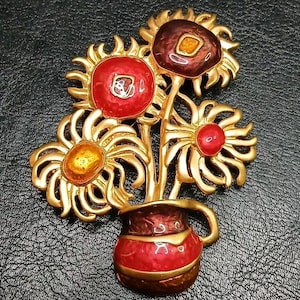 May include: A gold-tone metal brooch featuring a bouquet of three stylized flowers in a small pitcher. The flowers have vibrant red, orange, and deep maroon enamel centers with curved petals. The pitcher is finished in a glossy red enamel.