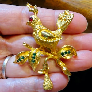 May include: A gold-tone brooch featuring a bird perched on a branch with green leaves and a dangling pear. The bird has a red rhinestone eye and a gold-tone body.