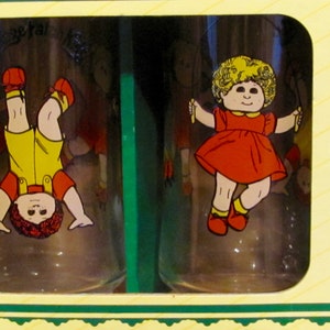May include: A set of two clear glass cups with a Cabbage Patch Kids theme. One cup features a girl doll with blonde hair wearing a red dress and holding jump ropes. The other cup features a boy doll with brown hair wearing a yellow shirt and red shorts, standing upside down.