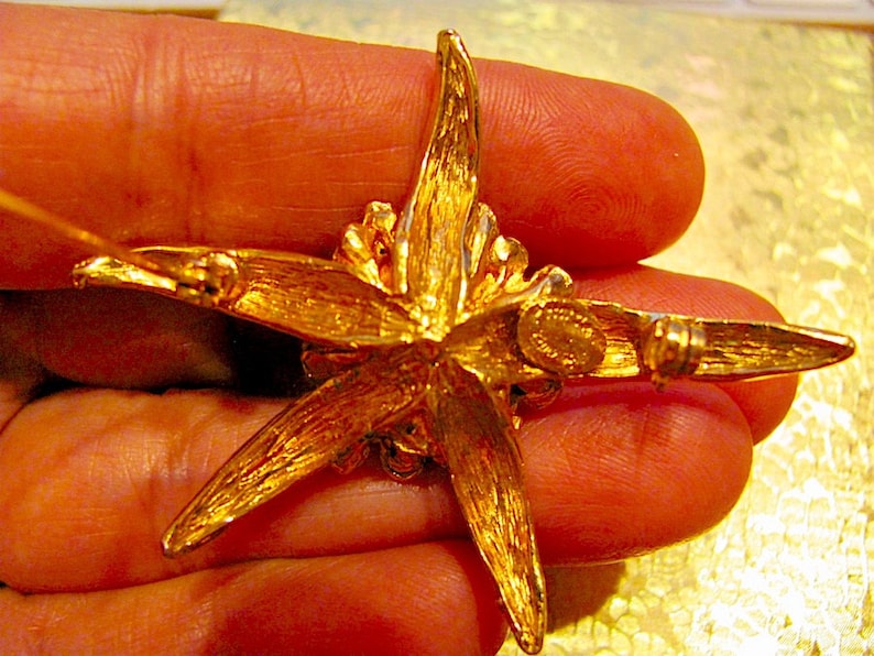 May include: A gold-toned starburst brooch with a textured surface. The brooch has five pointed rays and a central design. The back of the brooch features a pin clasp and a small, circular maker's mark.