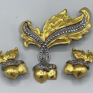 May include: A gold-toned jewelry set featuring a brooch and two matching earrings. The brooch is leaf-shaped with a silver-toned, crystal-studded accent. The earrings and brooch feature acorn designs with gold and silver tones.