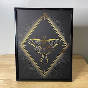 May include: A black framed print with a gold geometric design featuring a luna moth. The moth is outlined in gold and has a black background with gold glitter.