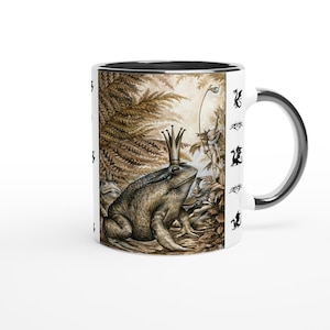 Puede incluir: Taza de cerámica blanca con interior y asa negros. La taza presenta una ilustración detallada de una rana con corona, rodeada de otras ranas y follaje. El exterior de la taza tiene un patrón repetido de figuras negras con forma de dragón.