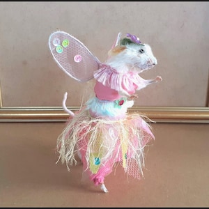 May include: A whimsical fairy mouse figurine with a white body, adorned with a pink tutu, wings, and a matching bodice. The mouse wears a small hat with a purple accent. The wings are embellished with colorful sequins, creating a charming, handcrafted aesthetic.