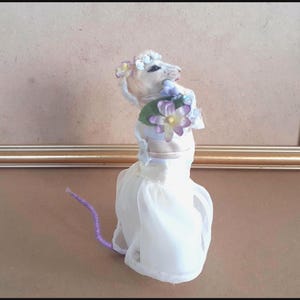 May include: A whimsical, handmade mouse figurine wearing a white dress and a floral headpiece. The mouse has a lavender tail and is posed against a neutral background, creating a charming, fairytale aesthetic.