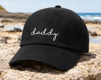 May include: Black baseball cap with the word "daddy" embroidered in white script. The cap has a curved brim and a button on top. The background shows a beach scene with rocks and water.