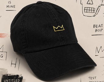 May include: Black baseball cap with a gold crown embroidered on the front. The cap is part of a collection with graffiti-style art and text, including "CROWN IS MINE" and "THE MOST KINGS GET THEIR HEAD CUT OFF."