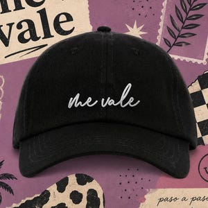 May include: Black baseball cap with the embroidered text "me vale" in white script. The hat has a curved brim and a rounded crown. The background features a collage of purple, white, and black elements, including text and illustrations.