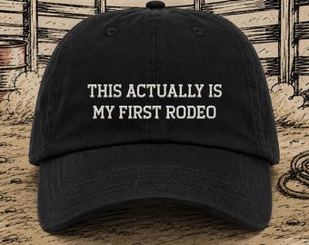 May include: Black baseball cap with the embroidered text "THIS ACTUALLY IS MY FIRST RODEO" in white. The cap has a curved brim and a rounded crown. The background features a rodeo-themed illustration.