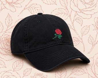May include: Black baseball cap with a curved brim. The cap features a red rose with green leaves embroidered on the front. The background is a light pink color with a rose pattern.