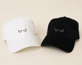 May include: Two baseball caps, one white and one black, are displayed. Each cap features a minimalist, black line drawing of breasts. The caps have a curved brim and an adjustable strap at the back, suitable for various head sizes.