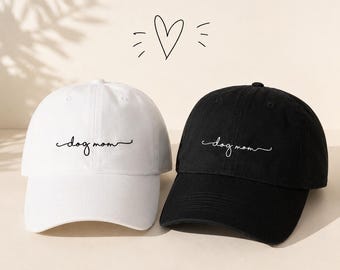 May include: A white and a black baseball cap. Each cap has the cursive text "dog mom" embroidered on the front. A simple heart and lines are drawn above the caps.