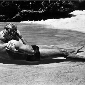 May include: A black and white photograph of a couple kissing on a beach. The man is wearing swimming trunks and the woman is wearing a swimsuit. The couple are lying on their sides, facing each other.