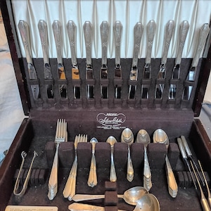 Puede incluir: Un estuche de madera que contiene un juego de cubiertos de plata. El set incluye doce cuchillos con mangos decorados, varios tenedores, diversas cucharas, un cucharón y utensilios de servicio sobre un forro de tela marrón oscuro.