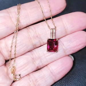 Puede incluir: Un collar dorado con un colgante rectangular rojo oscuro. El colgante está adornado con pequeñas piedras transparentes. La cadena es delicada y tiene un cierre de mosquetón. El collar se muestra en una mano.