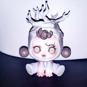 May include: A small figurine featuring a pale, round-faced character with large, light-colored eyes and a dark, branch-like marking above the left eye. The figure wears a white, hood-like headpiece topped with white, tree-branch-like structures.