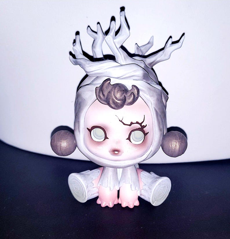 May include: A small figurine featuring a pale, round-faced character with large, light-colored eyes and a dark, branch-like marking above the left eye. The figure wears a white, hood-like headpiece topped with white, tree-branch-like structures.