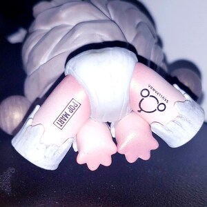 May include: A collectible figurine featuring a grey textured upper body and pink legs wearing a white diaper. The left leg is printed with the text POP MART in a rectangular box, and the right leg features the text SKULLPANDA with a circular logo.