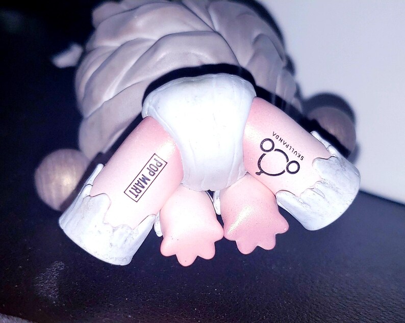 May include: A collectible figurine featuring a grey textured upper body and pink legs wearing a white diaper. The left leg is printed with the text POP MART in a rectangular box, and the right leg features the text SKULLPANDA with a circular logo.
