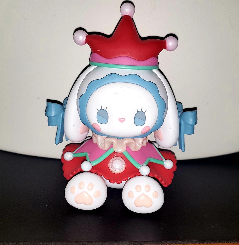May include: A white rabbit figurine wearing a red jester hat with pink spheres, a blue hood, and a red dress with green trim. The figure has long white ears with blue bows, a ruffled collar, and white feet featuring light pink paw pad details.