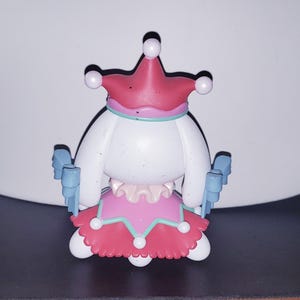May include: A small figurine featuring a white character wearing a pink jester-style hat with white spheres at the tips, a pink skirt with a teal trim and white spheres, and light blue bow-shaped accents on the sides of the body.