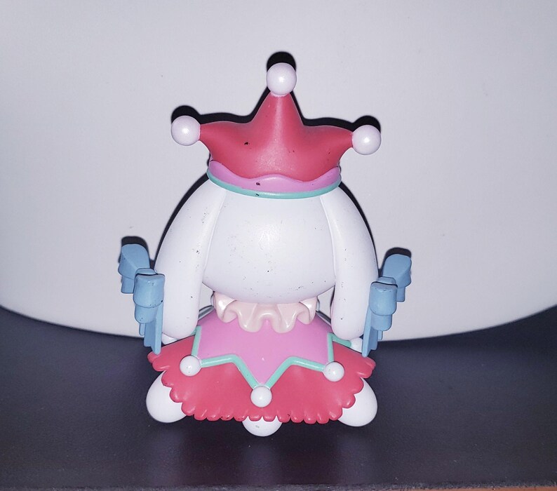 May include: A small figurine featuring a white character wearing a pink jester-style hat with white spheres at the tips, a pink skirt with a teal trim and white spheres, and light blue bow-shaped accents on the sides of the body.