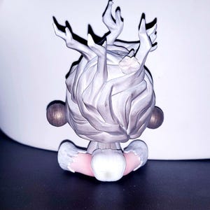 May include: A small figurine featuring a stylized head with light gray, textured hair that resembles branching antlers or tree limbs. The figure has two round, dark brown spheres on either side and a pale pink neck area with white trim.