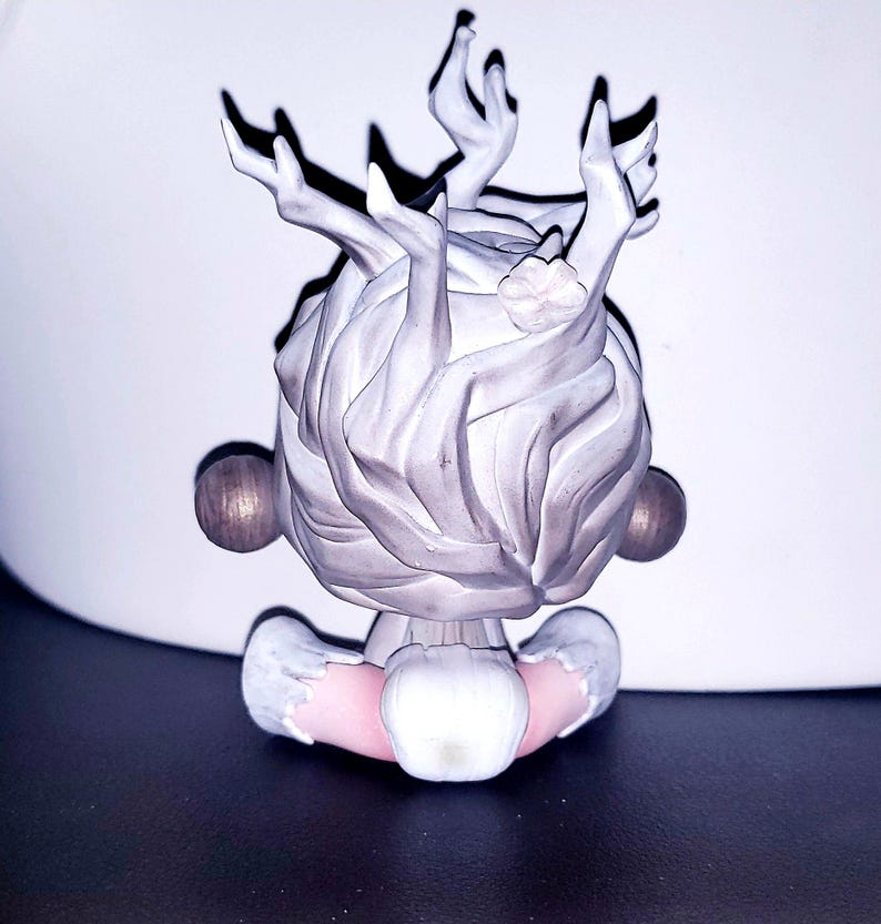 May include: A small figurine featuring a stylized head with light gray, textured hair that resembles branching antlers or tree limbs. The figure has two round, dark brown spheres on either side and a pale pink neck area with white trim.