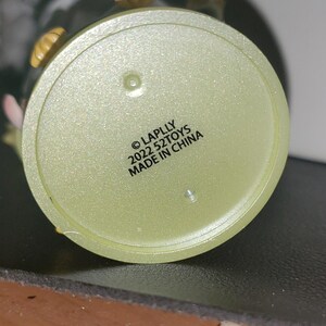 May include: A circular, pale yellow, translucent plastic base for a collectible figure. The underside features black printed text that reads: &copy; LAPLLY 2022 52TOYS MADE IN CHINA. The surface has a subtle glittery texture and two small circular indentations.