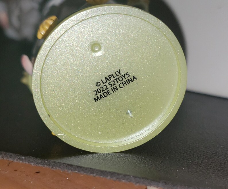 May include: A circular, pale yellow, translucent plastic base for a collectible figure. The underside features black printed text that reads: &copy; LAPLLY 2022 52TOYS MADE IN CHINA. The surface has a subtle glittery texture and two small circular indentations.