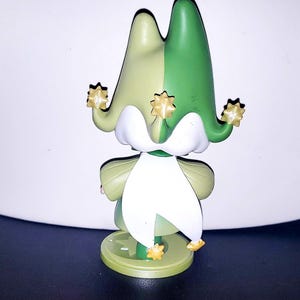 May include: A small figurine featuring a jester-style hat split into light green and dark green sections. The hat has three points, each tipped with a small gold star. A white collar drapes over the shoulders, and the figure stands on a round base.
