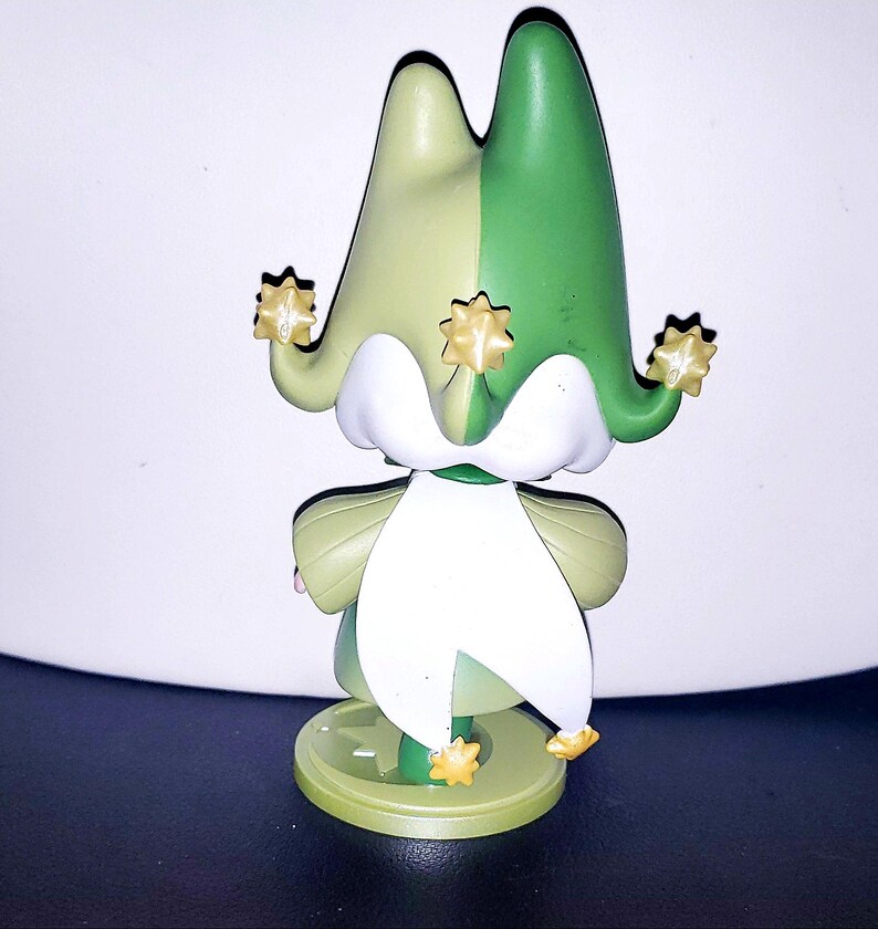 May include: A small figurine featuring a jester-style hat split into light green and dark green sections. The hat has three points, each tipped with a small gold star. A white collar drapes over the shoulders, and the figure stands on a round base.