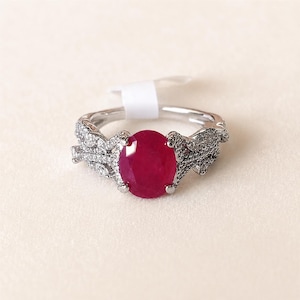 May include: A silver-toned ring featuring a large, oval-cut deep red gemstone at the center. The band is intricately designed with a vine-like pattern encrusted with numerous small, clear, sparkling stones, and includes a small white price tag.
