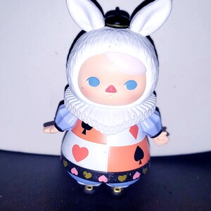 May include: A small figurine of a character wearing a white rabbit-eared hood, a ruffled collar, and a dress featuring a checkered pattern with red hearts, red diamonds, and black spades, accented by a black band with small pink and gold hearts.