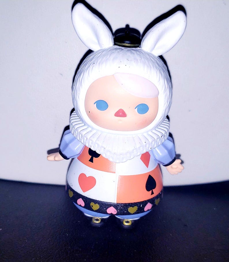 May include: A small figurine of a character wearing a white rabbit-eared hood, a ruffled collar, and a dress featuring a checkered pattern with red hearts, red diamonds, and black spades, accented by a black band with small pink and gold hearts.