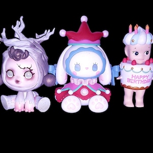 May include: Five collectible vinyl figures arranged in a row. The first is a green and white jester-like character, the second is a pale purple creature with branch-like antlers, the third is a white rabbit-eared figure in a red and blue outfit, the fourth is a figure wearing a cake costume with the text "HAPPY BIRTHDAY", and the fifth is a white rabbit-eared figure in a playing card patterned outfit.
