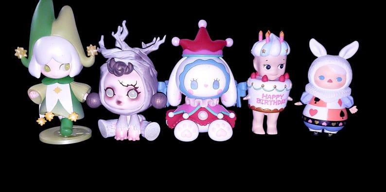 May include: Five collectible vinyl figures arranged in a row. The first is a green and white jester-like character, the second is a pale purple creature with branch-like antlers, the third is a white rabbit-eared figure in a red and blue outfit, the fourth is a figure wearing a cake costume with the text "HAPPY BIRTHDAY", and the fifth is a white rabbit-eared figure in a playing card patterned outfit.